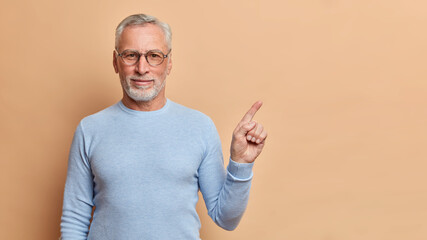 I recommend you to use this copy space. Self confident grey haired European man points at upper right corner shows great place dressed in casual blue jumper isolated over brown studio background