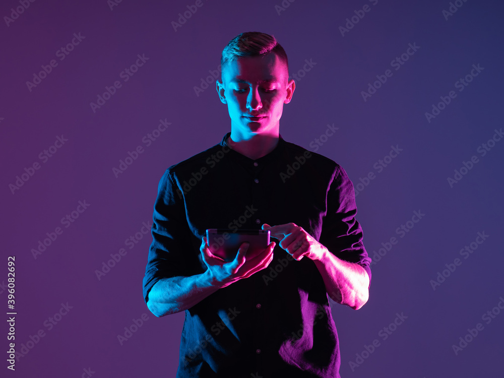 Man with a tablet on a purple background. It is illuminated by neon light. Concept - he uses a tablet application. Guy is using the app in the gadget. Student is using some kind of application - obrazy, fototapety, plakaty 