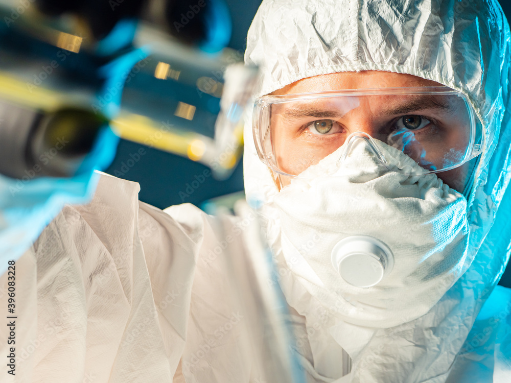 Man in a biological protection suit close-up. He is holding test tube ...