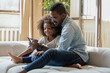 © fizkes - Let us call mommy. Happy african black family caring young father and smiling preteen daughter relaxing on couch hugging looking on phone screen making answering videocall, watching funny photo online