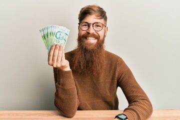 Sticker - Young irish redhead man holding 5 singapore dollars banknotes sitting on the table looking positive and happy standing and smiling with a confident smile showing teeth