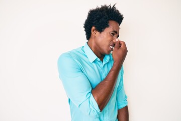 Sticker - Handsome african american man with afro hair wearing casual clothes smelling something stinky and disgusting, intolerable smell, holding breath with fingers on nose. bad smell