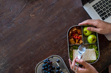 Close up of man hands at desk with a laptop at home or in office with healthy vegan meal in reusable stainless steel lunch box. Healthy lifestyle, zero waste and sustainable plastic free lifestyle