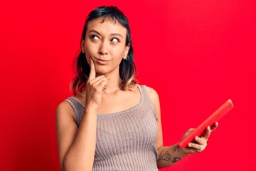 Wall Mural - Young woman holding touchpad tablet serious face thinking about question with hand on chin, thoughtful about confusing idea