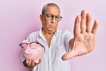 Sticker - Handsome mature senior man holding piggy bank with glasses with open hand doing stop sign with serious and confident expression, defense gesture