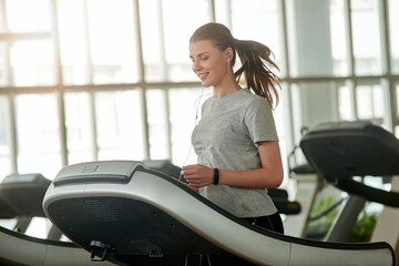 Wall Mural - Young beautiful lady running on treadmill. Pretty smiling female person exercising at gym. Youth, sport, active way of life.