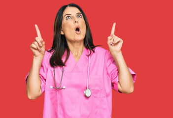 Poster - Middle age brunette woman wearing doctor uniform and stethoscope amazed and surprised looking up and pointing with fingers and raised arms.