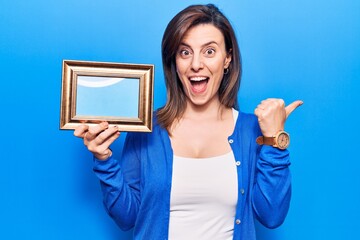 Wall Mural - Young beautiful woman holding empty frame pointing thumb up to the side smiling happy with open mouth