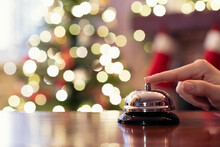 Hand Of Guest Ringing Reception Bell On Desk Of Guesthouse, Hotel At Christmas Time. Color Shining Garland On Christmas Tree On Background. Travel Concept.