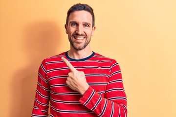 Canvas Print - Young handsome man wearing casual striped t-shirt standing over isolated yellow background smiling cheerful pointing with hand and finger up to the side