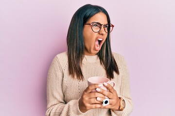 Poster - Young latin girl holding cup of tea angry and mad screaming frustrated and furious, shouting with anger. rage and aggressive concept.