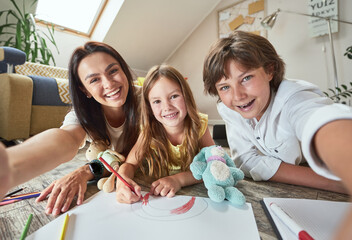 Wall Mural - Young cheerful mother with two happy children lying on the floor in the living room and making family selfie while spending time together at home