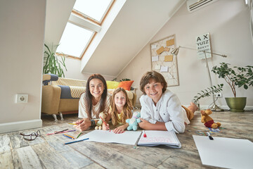 Wall Mural - Happy caucasian family lying on the floor in living room at home and smiling at camera, mother spending time with children