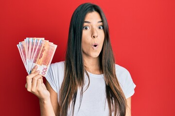 Sticker - Young brunette woman holding 100 norwegian krone banknotes scared and amazed with open mouth for surprise, disbelief face