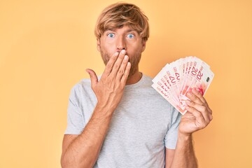 Poster - Young blond man holding israeli shekels banknotes covering mouth with hand, shocked and afraid for mistake. surprised expression