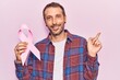Young handsome man holding pink cancer ribbon smiling happy pointing with hand and finger to the side