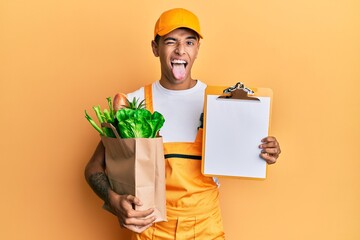 Sticker - Young handsome african american man wearing courier uniform with groceries from supermarket and clipboard sticking tongue out happy with funny expression.