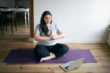 Calm mindful barefoot young female with curvy body practicing meditation at home, sitting in padmasana with eyes closed, holding hands pressed together in namaste, doing breathing exercises