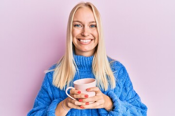 Poster - Young blonde girl wearing winter sweater and drinking a cup of hot coffee smiling with a happy and cool smile on face. showing teeth.