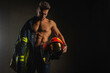 young handsome adult, muscular firefighter in uniform holding ax of fire equipment in his hands, pensive, isolated on dark background. Low key. Protection concept. there is a place for an inscription