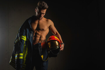 Wall Mural - young handsome adult, muscular firefighter in uniform holding ax of fire equipment in his hands, pensive, isolated on dark background. Low key. Protection concept. there is a place for an inscription