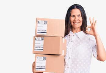 Young beautiful brunette woman holding delivery cardboard package doing ok sign with fingers, smiling friendly gesturing excellent symbol