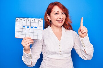 Sticker - Young redhead woman holding weather calendar showing rainy week over blue background smiling with an idea or question pointing finger with happy face, number one