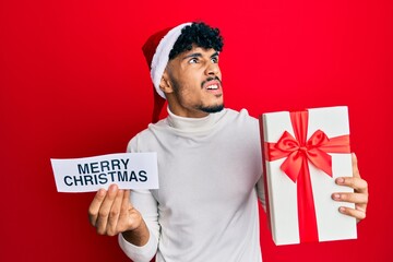 Young arab handsome man wearing christmas hat holding merry christmas message angry and mad screaming frustrated and furious, shouting with anger looking up.