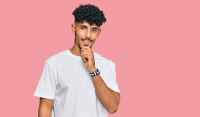 Young arab man wearing casual white t shirt looking confident at the camera smiling with crossed arms and hand raised on chin. thinking positive.