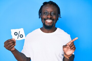 Wall Mural - Young african american man with braids holding percentage symbol paper smiling happy pointing with hand and finger to the side