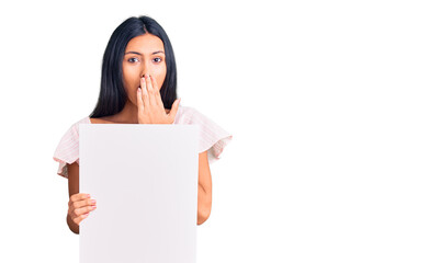 Wall Mural - Young beautiful latin girl holding blank empty banner covering mouth with hand, shocked and afraid for mistake. surprised expression