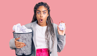 Canvas Print - Young african american woman with braids holding paper bin full of crumpled papers scared and amazed with open mouth for surprise, disbelief face