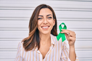 Young hispanic woman smiling happy holding awareness green ribbon standing at the city.