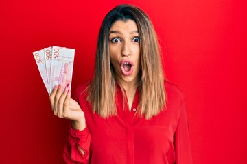 Sticker - Beautiful brunette woman holding 500 swedish krona banknotes scared and amazed with open mouth for surprise, disbelief face