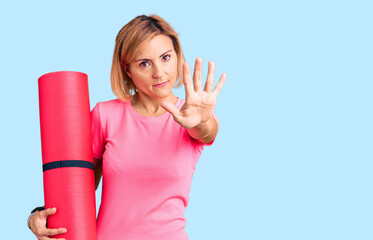 Sticker - Young blonde woman holding yoga mat with open hand doing stop sign with serious and confident expression, defense gesture