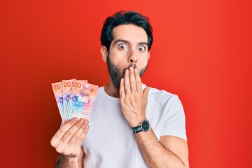 Canvas Print - Young hispanic man holding swiss franc banknotes covering mouth with hand, shocked and afraid for mistake. surprised expression