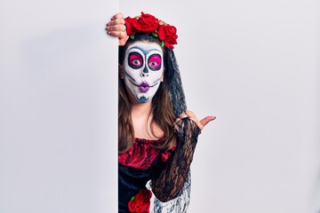 Wall Mural - Young woman wearing day of the dead custome holding blank empty banner surprised pointing with hand finger to the side, open mouth amazed expression.