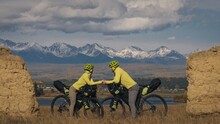 The Man And Woman Travel On Mixed Terrain Cycle Touring With Bikepacking. The Two People Journey With Bicycle Bags. Mountain Snow Capped, Stone Arch.