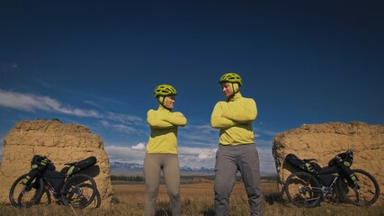 The man and woman travel on mixed terrain cycle touring with bikepacking. The two people journey with bicycle bags. Mountain snow capped, stone arch.