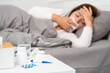 © Pichsakul - pill and glasses of water on the table woman suffering headache, fever and flu lying in bed background.