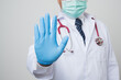 © Chanakon - Close Up Asian bald Doctor in white coat, stethoscope, blue glove wearing face mask showing rejecting gesture by stop palm sign in front of camera isolated on white background.