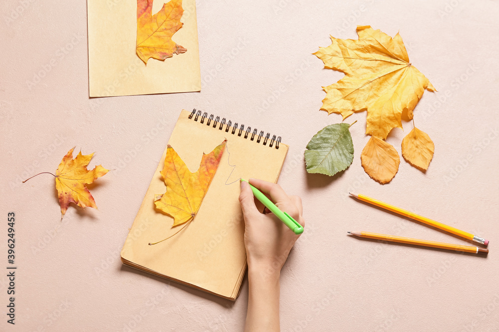 Woman drawing autumn leaf in notebook on color background