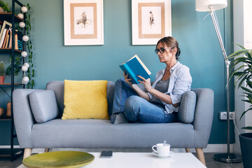 Mature beautiful woman reading a book sitting on couch at home.