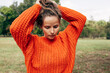 © iuricazac - The young woman in an orange knitted sweater arranges her hair in a ponytail against a nature background. The gorgeous female has a serious expression, resting outdoors in the park.