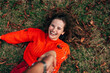 © iuricazac - Top view of a gorgeous young woman smiling broadly in orange knitted sweater lying on the grass with autumn leaves and taking the hand to follow. The beautiful female has a joyful expression outdoors.