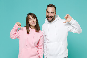 Smiling cheerful young couple two friends man woman 20s wearing white pink casual hoodie pointing thumbs on themselves looking camera isolated on blue turquoise colour wall background studio portrait.