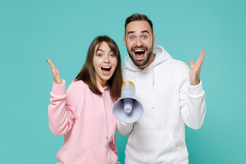 Canvas Print - Excited cheerful young couple two friends man woman 20s wearing white pink casual hoodie screaming in megaphone spreading hands isolated on blue turquoise colour wall background studio portrait.