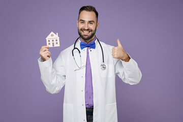 Smiling young bearded doctor man wearing white medical gown stethoscope hold house showing thumb up isolated on violet colour background studio portrait. Healthcare personnel health medicine concept.