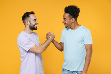 Wall Mural - Cheerful young friends european african american men 20s wearing casual violet blue t-shirts greeting holding hands looking at each other isolated on bright yellow colour background studio portrait.