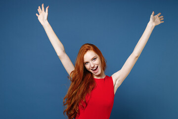 Wall Mural - Excited cheerful happy laughing young redhead woman 20s wearing bright red elegant evening dress standing rising spreading hands looking camera isolated on blue color wall background studio portrait.